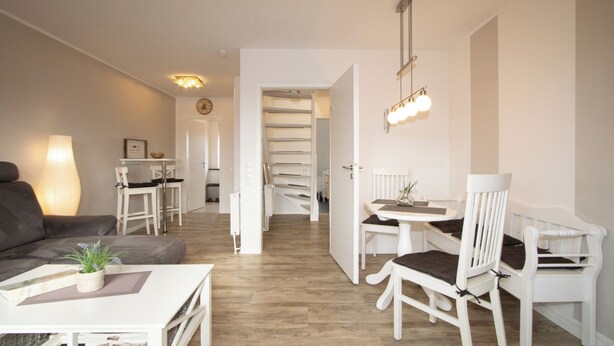 A modern living room with a sofa, a dining table with chairs, a staircase, and a chandelier. - TOP vacation apartment for up to 2 people. Bright and modern furnishings. (Hörnum)