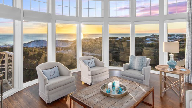 Habitación con amplios ventanales que ofrecen vistas al mar y colinas al atardecer. Hay dos sillones y una mesa de centro de madera con objetos decorativos.