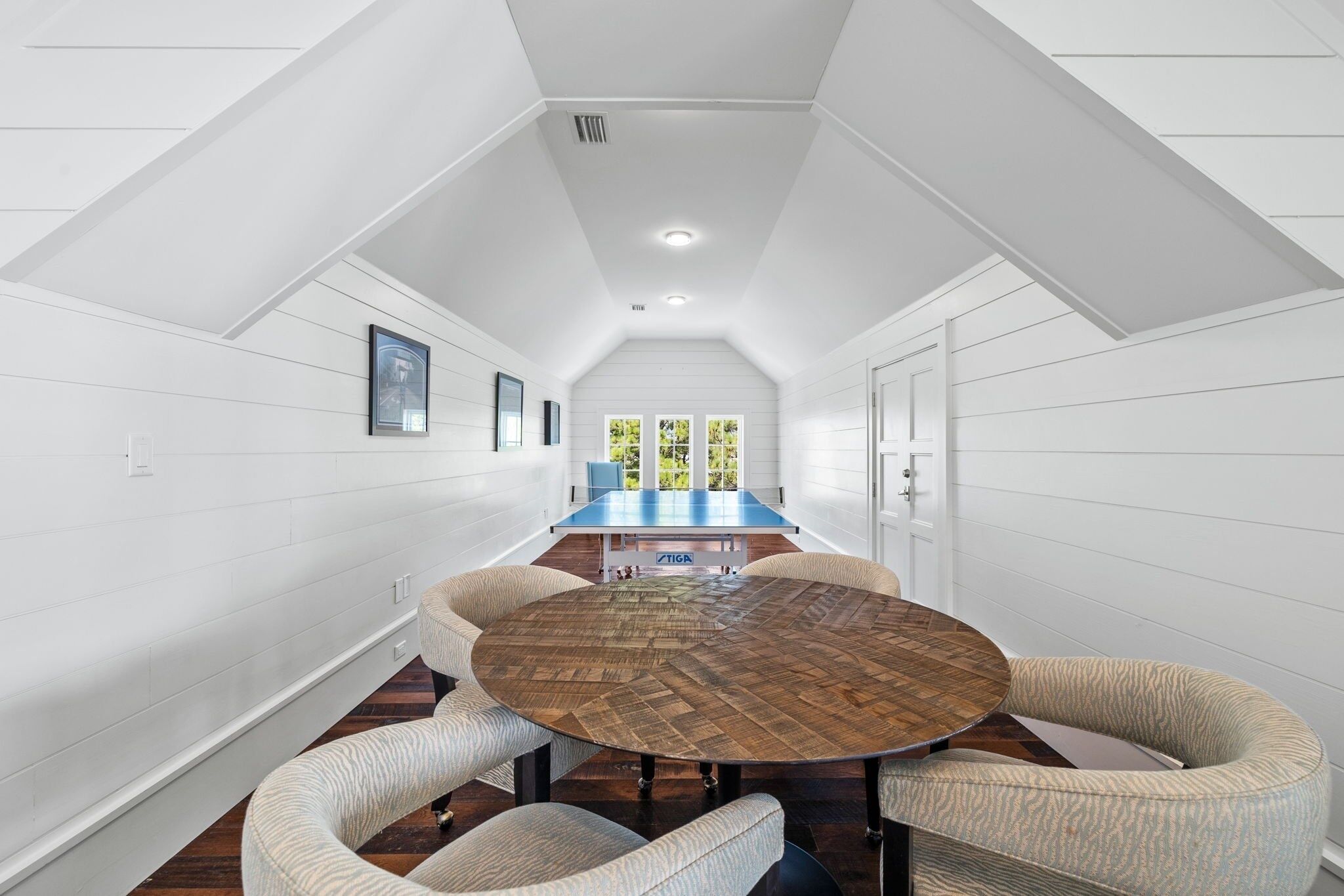 A spacious room with a round wooden table surrounded by cushioned chairs, a blue sofa, and a large window with a view of greenery.