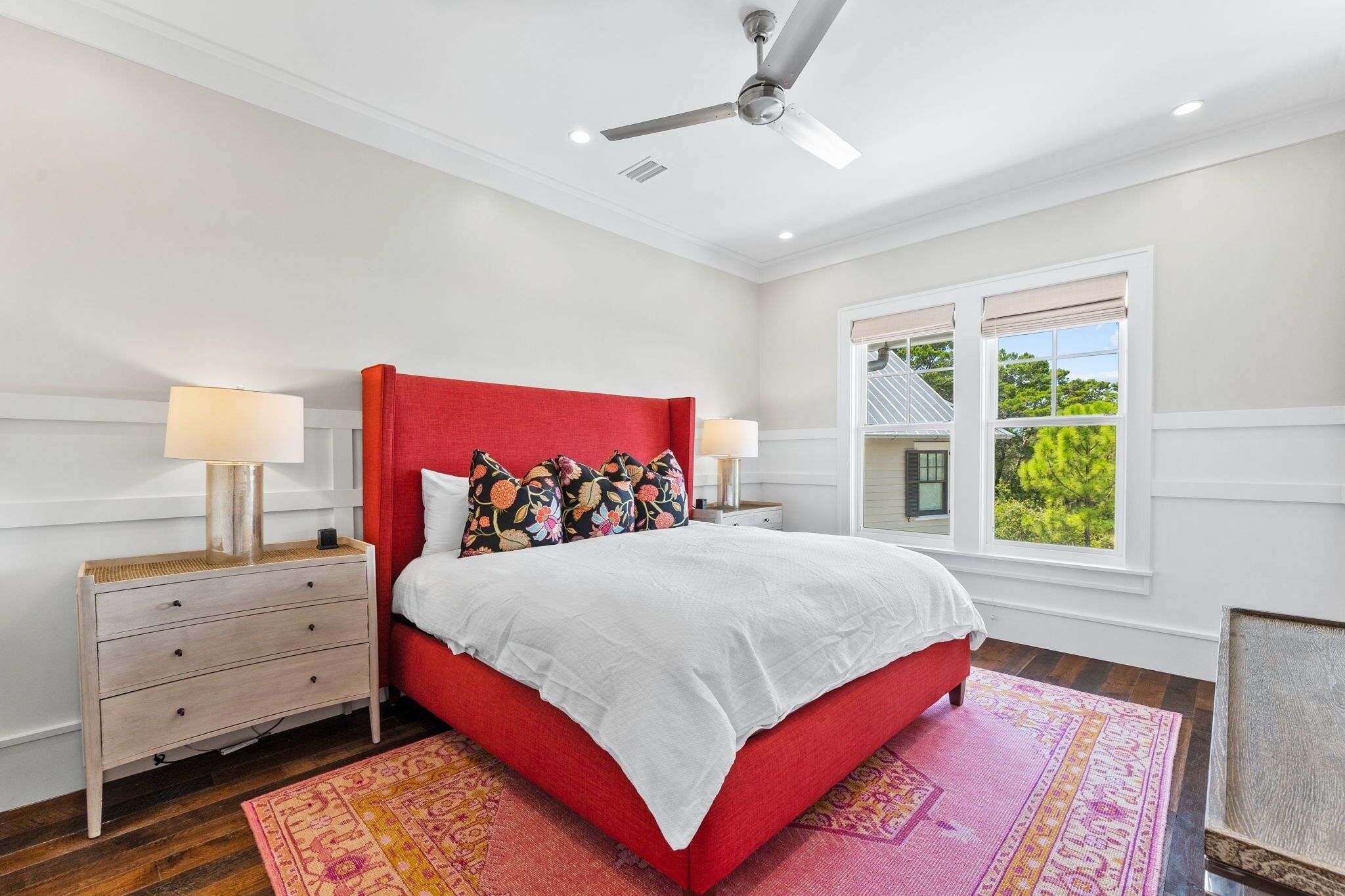 A bedroom with a red bed, white walls, a ceiling fan, and a window with a view of greenery.