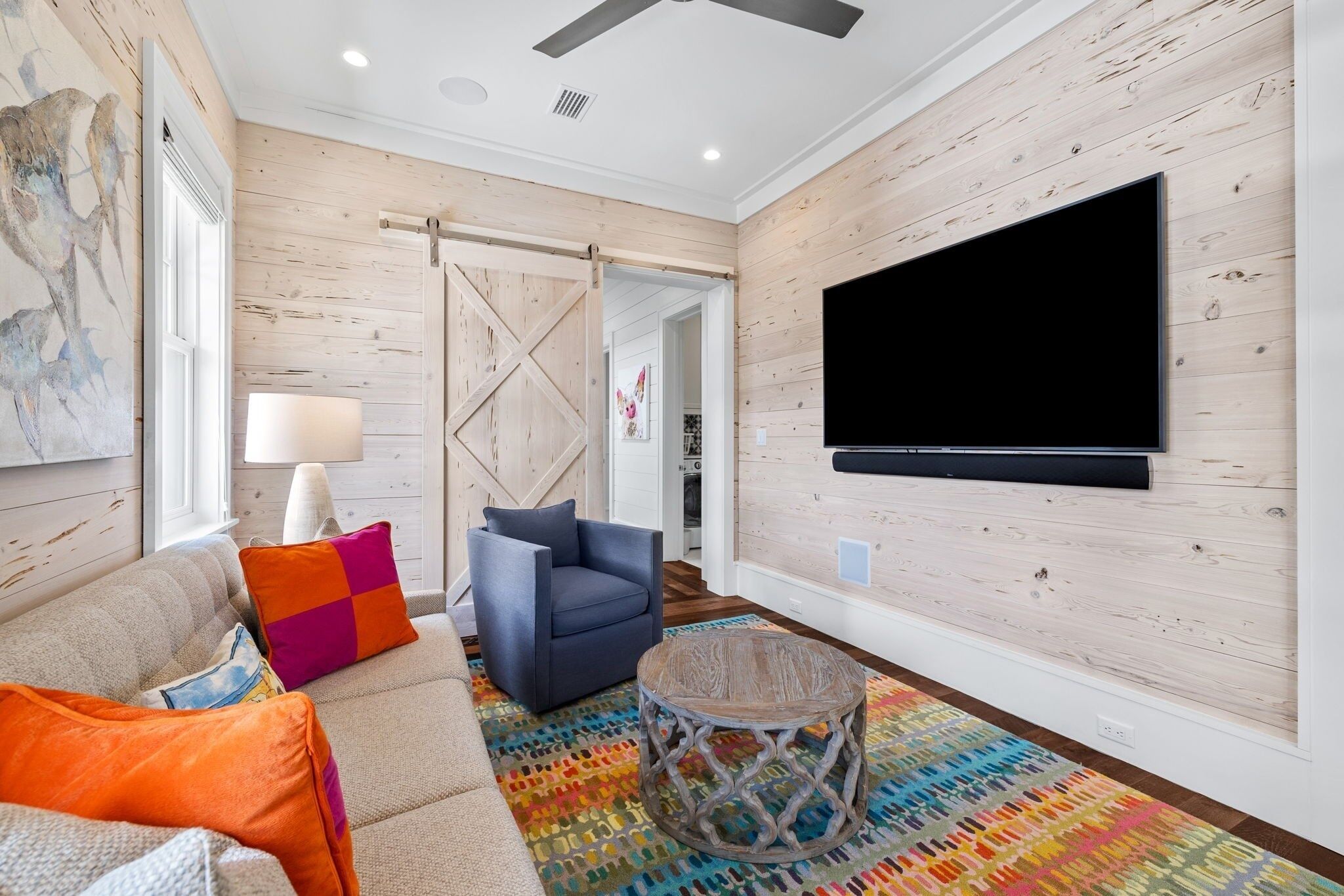 A modern living room with a large flat-screen TV, a blue armchair, a colorful rug, and a wooden ceiling fan.
