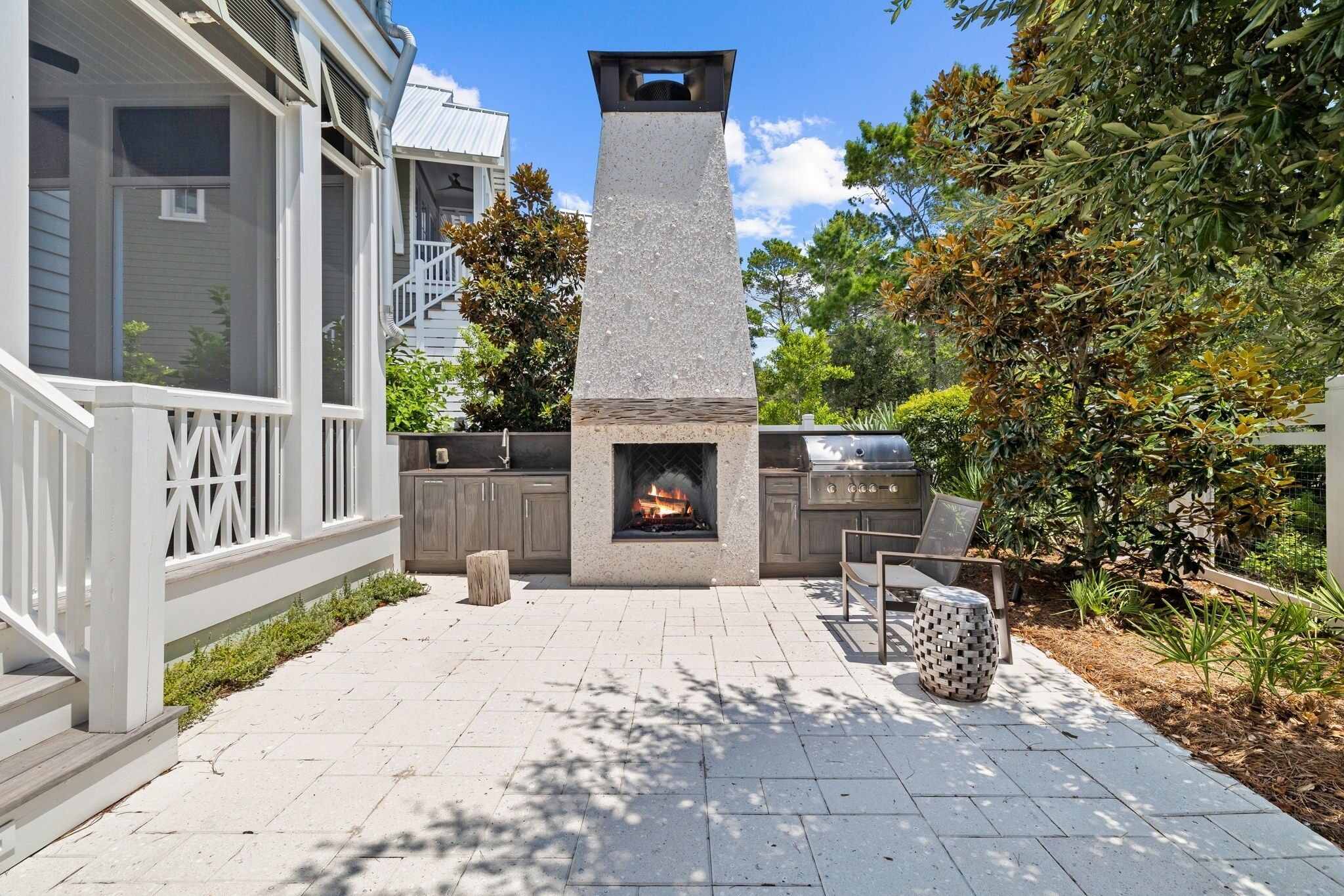 A stone fireplace with a fire, surrounded by a patio with a wooden fence, outdoor seating, and a view of a house with a balcony.