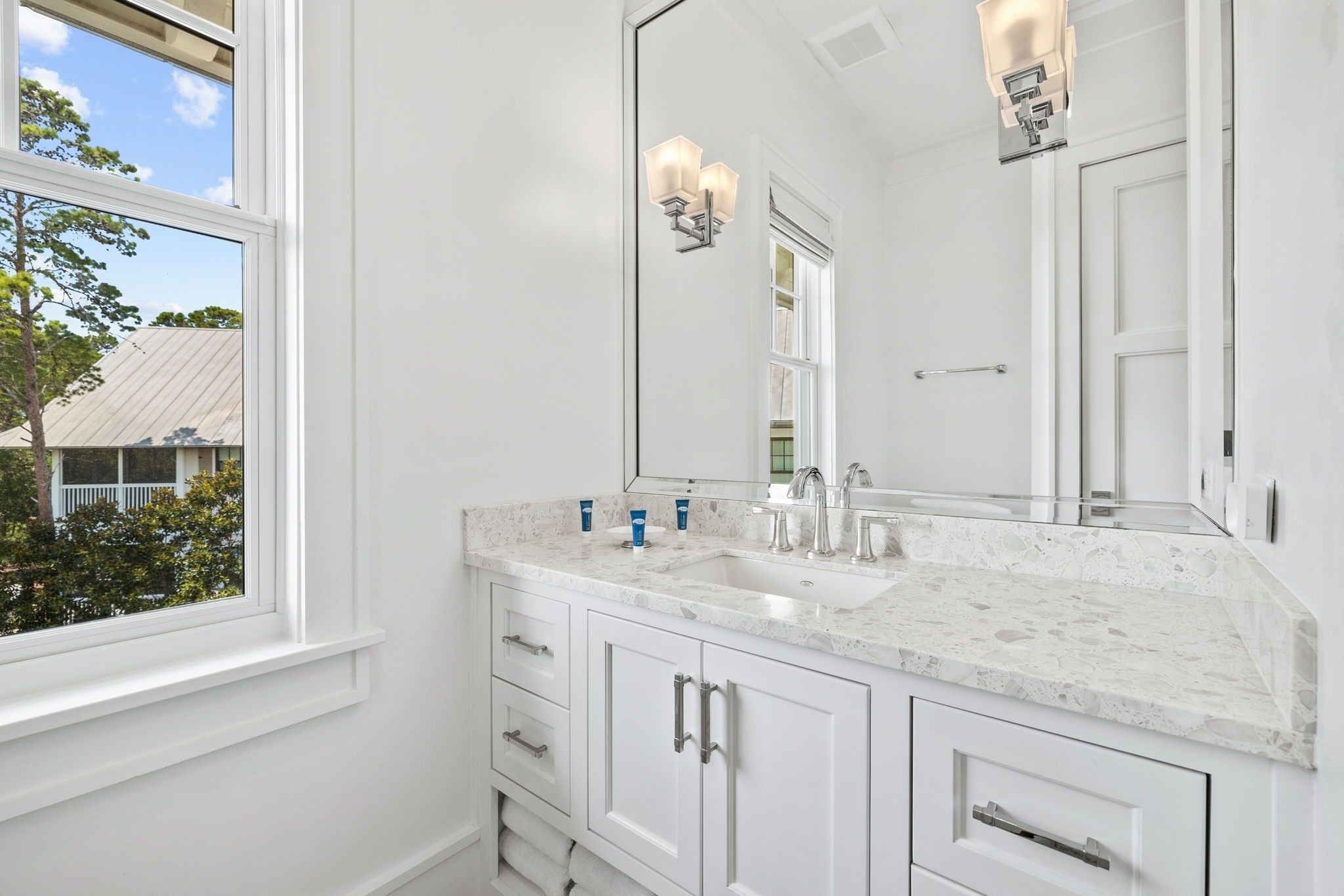 A bathroom with a large mirror, white cabinetry, a double sink, and a window with a view of a house and trees.