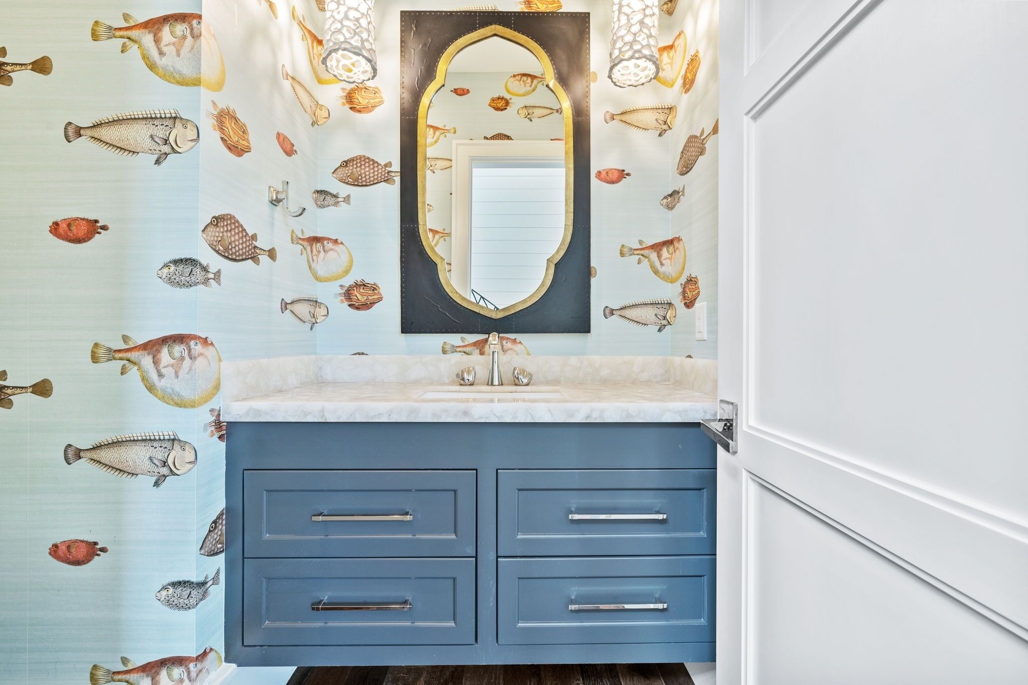 Een badkamer met een blauwe wastafel, een spiegel met gouden lijst en behang met een vismotief.