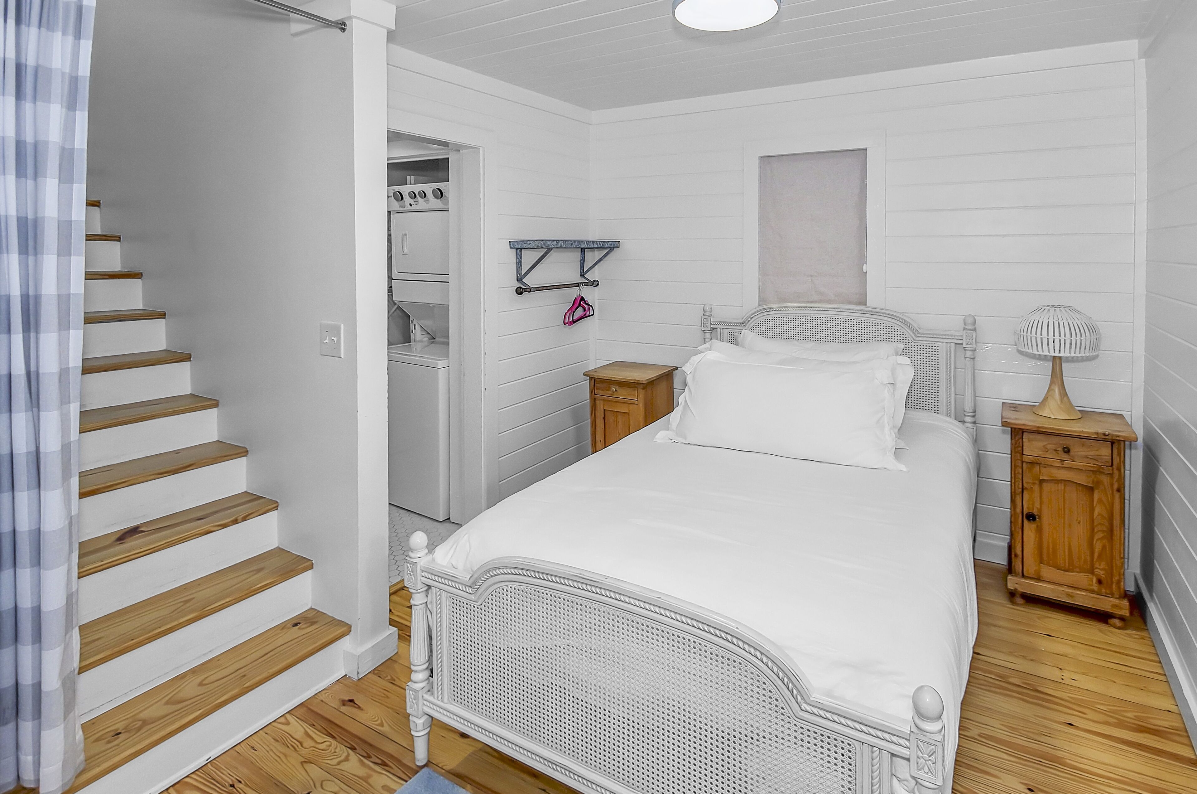 A bedroom with a white bed, wooden flooring, a wooden nightstand, a small wooden cabinet, a white ceiling, and a staircase.