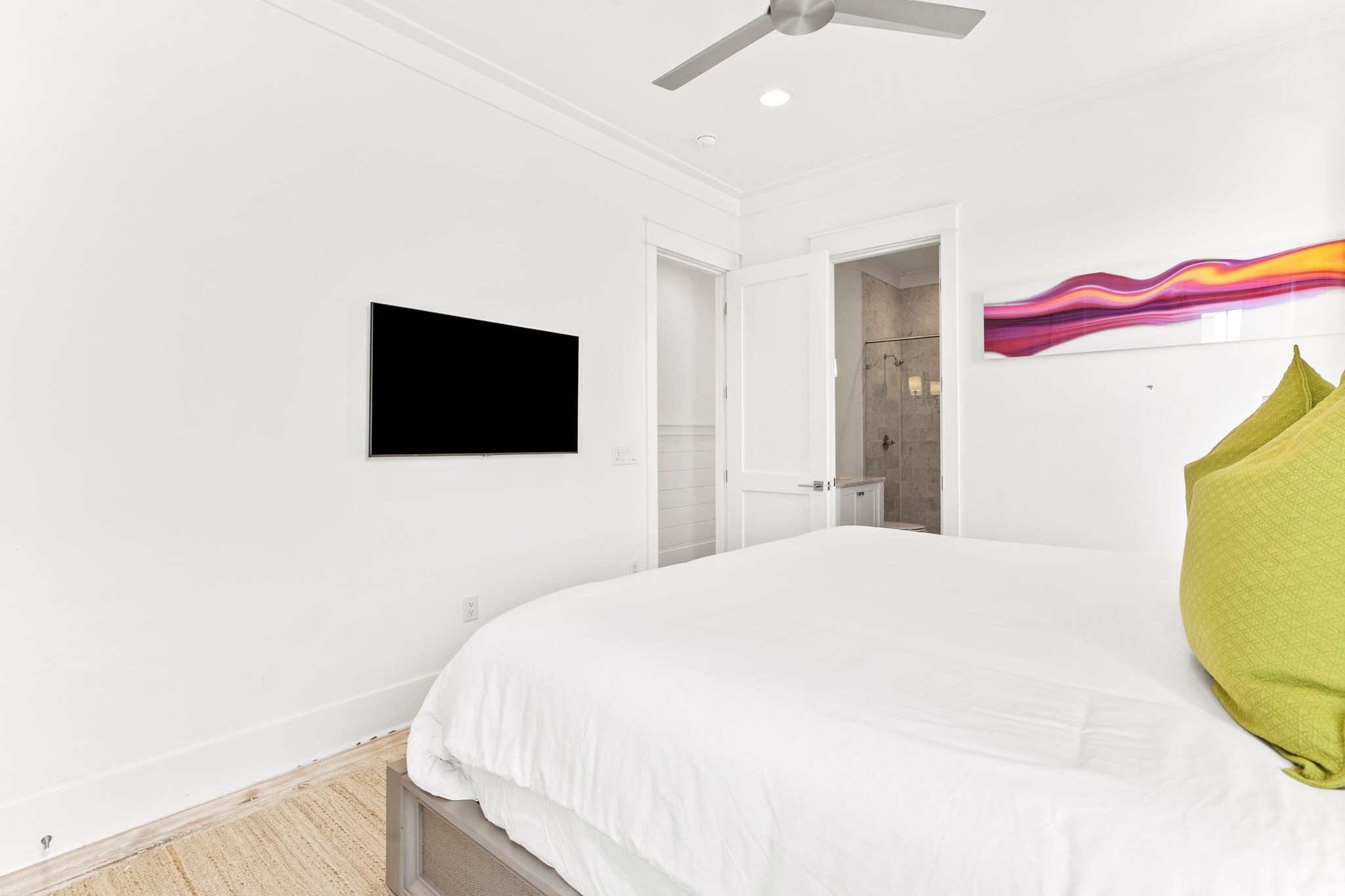 Quarto com cama, ventilador de teto, televisão de tela plana, porta de acesso a um banheiro e um quadro abstrato colorido na parede.