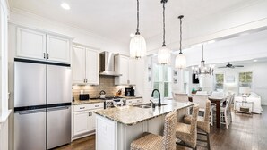 A modern kitchen with white cabinetry, a stainless steel refrigerator, a granite countertop, and a bar area with wicker chairs. - 'seas The Day' In Watercolor Beach 5 Bdrm/slps 10 Steps To Dragonfly Pool! 5 Bedroom Home by RedAwning (Santa Rosa Beach)