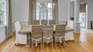 Uma sala de jantar com mesa de madeira e cadeiras de vime, um candelabro e janelas grandes com cortinas.