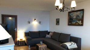 A modern living room with a grey sectional sofa, a wooden coffee table, and a white table lamp. There is a painting on the wall and a small rug on the floor.