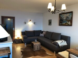 Un salon moderne avec un canapé d’angle gris, une table basse en bois et une lampe de table blanche. Un tableau est accroché au mur et un petit tapis recouvre le sol.