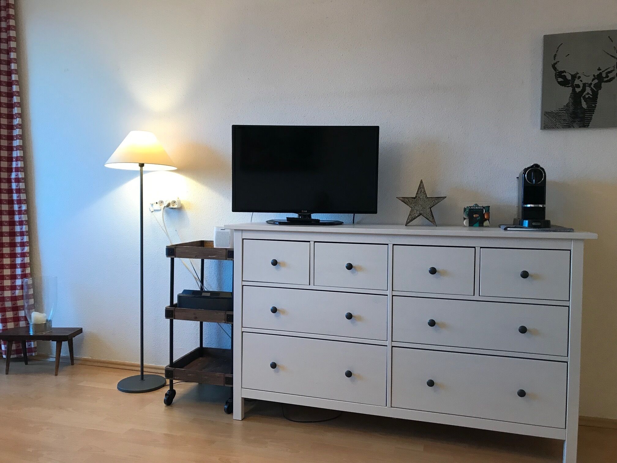 A white dresser with drawers, a flat-screen TV on top, a star decoration, a lamp, and a shelf with a coffee maker.