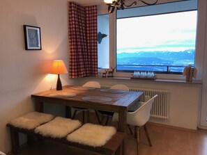 Un coin repas avec une table en bois, des chaises blanches et une vue sur les montagnes par la fenêtre.