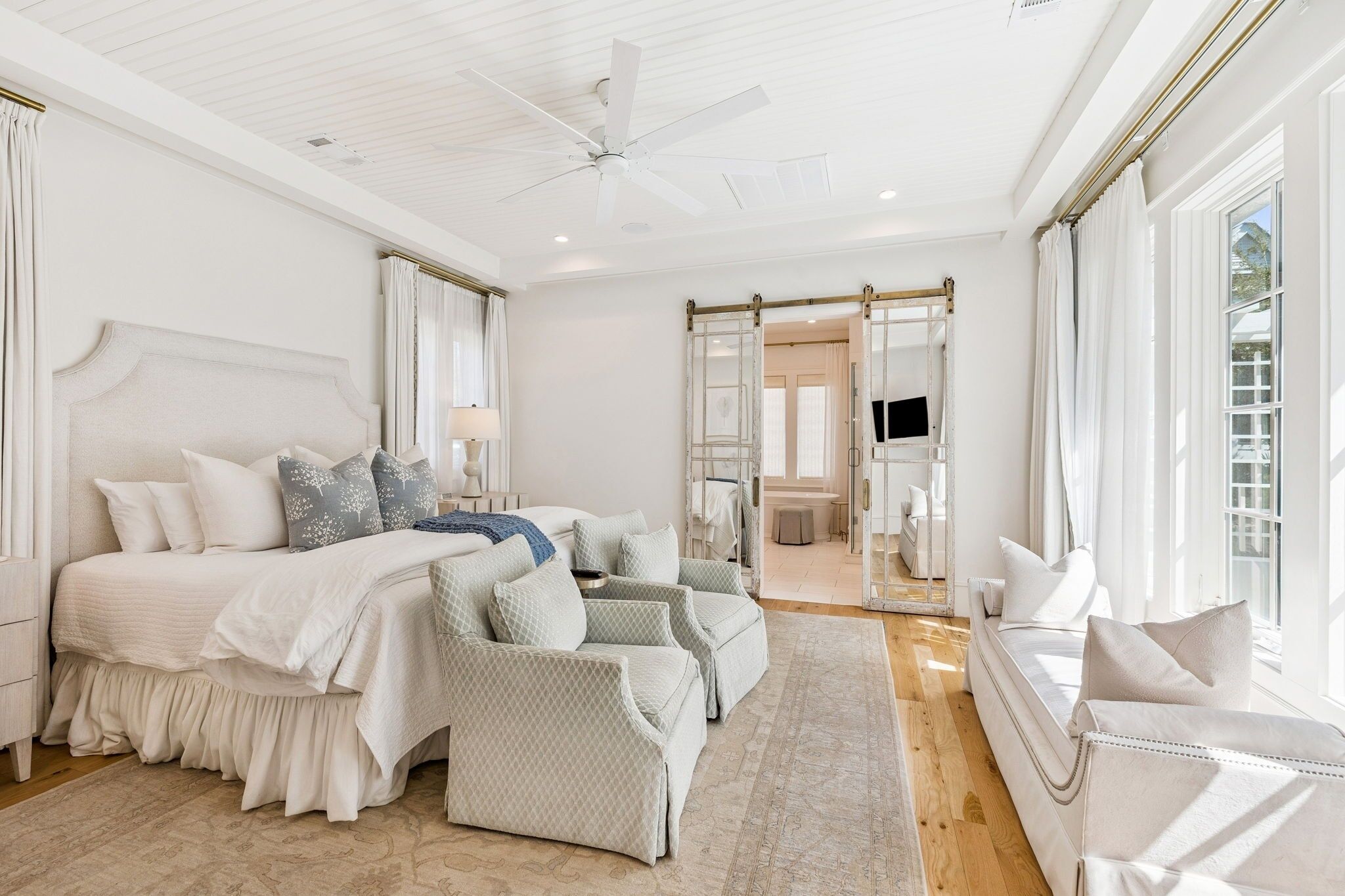 A bedroom with a large bed, two armchairs, a sofa, a ceiling fan, and a sliding glass door leading to a bathroom.