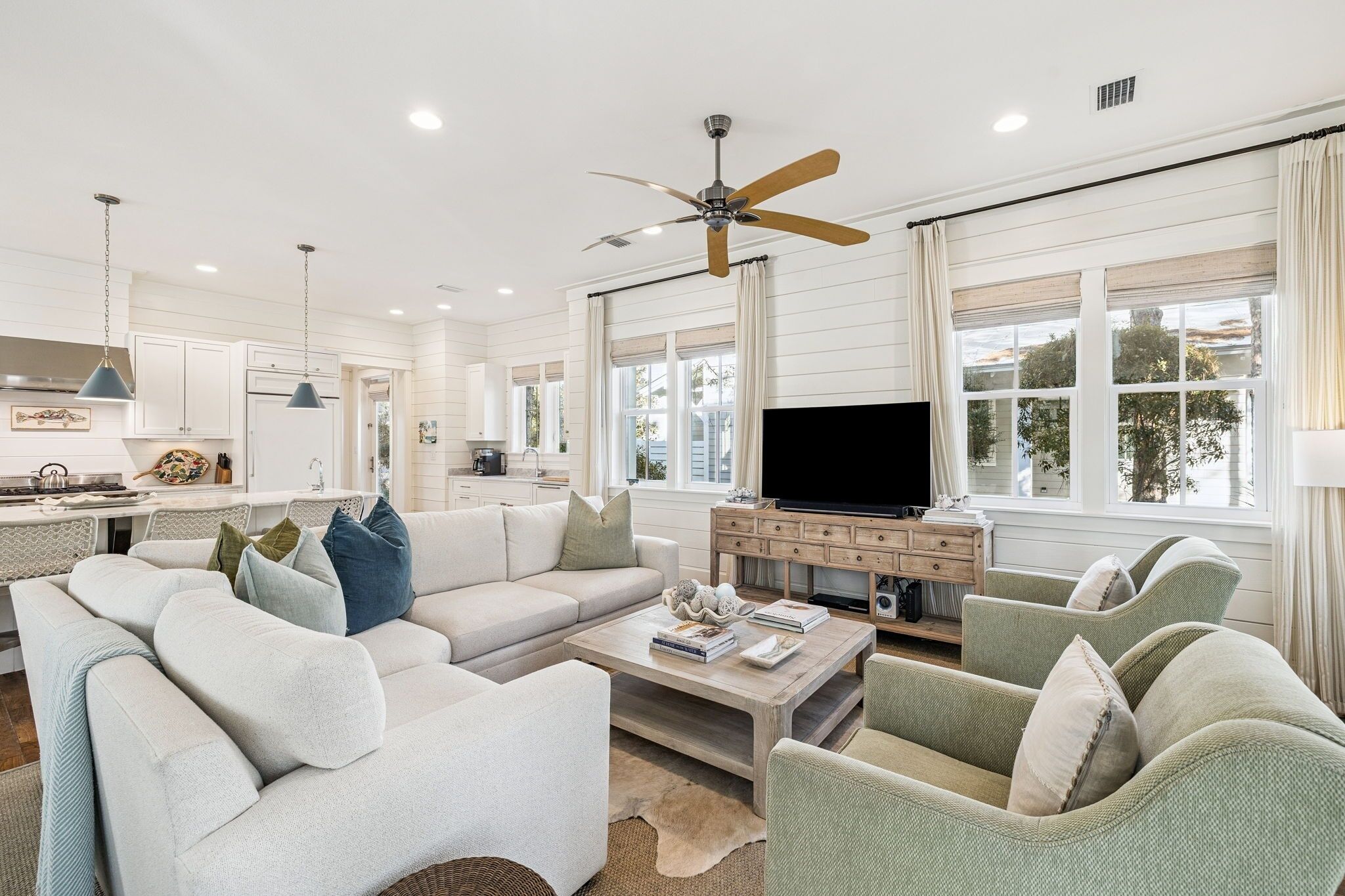 A spacious living room with a large sofa, two armchairs, a coffee table, and a TV. There is a ceiling fan, a kitchen area with white cabinets, and large windows with curtains.