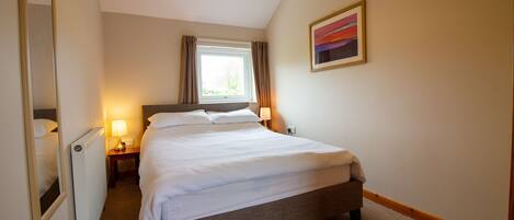 A neatly made bed in a hotel room with a window, a bedside lamp, and a framed picture on the wall.