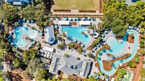 Eine Vogelperspektive zeigt ein Resort mit mehreren Schwimmbecken, Liegestühlen und einem Gebäude mit geneigtem Dach.
