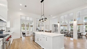 A spacious kitchen with white cabinetry, a large island, and a dining area with wooden tables and chairs. - Located Across From Camp Watercolor Luxurious 5br/7bth Home + Golf Cart 6 Bedroom Home by RedAwning (Santa Rosa Beach)