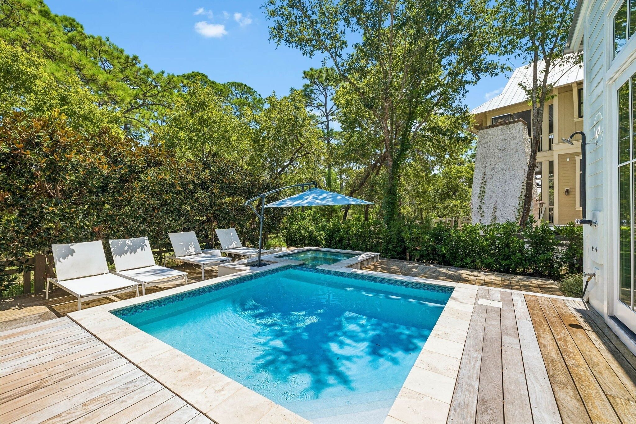 A rectangular swimming pool with a wooden deck and lounge chairs, surrounded by lush greenery and trees.
