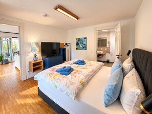 Une chambre moderne avec un grand lit, une télévision et une salle de bain visible à travers une porte ouverte.