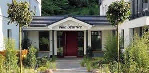 A building named Villa Beatrice with a red door, surrounded by greenery and a pathway leading to the entrance.