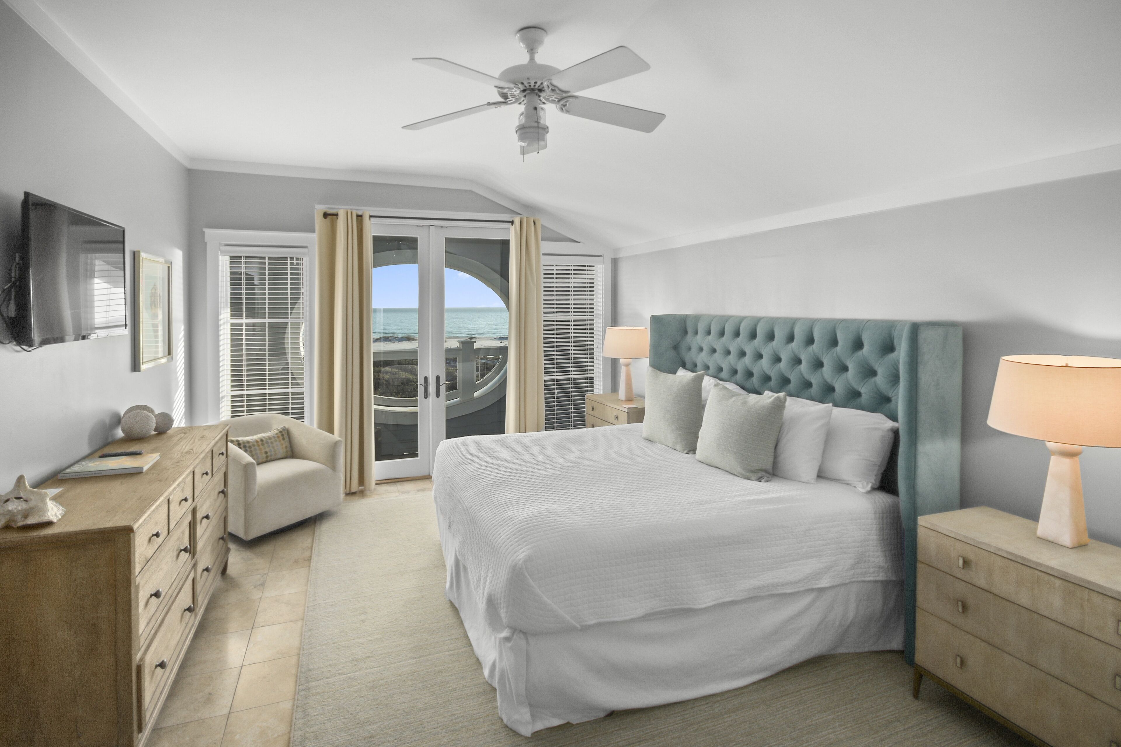 Une chambre avec un grand lit, un ventilateur de plafond, une table de chevet, une commode, une chaise et une vue sur l’océan par une porte coulissante.