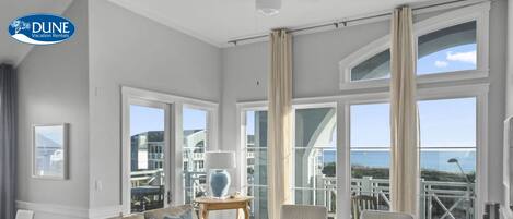 A spacious living room with a large window overlooking the ocean, furnished with a beige sofa, white armchairs, and a coffee table.