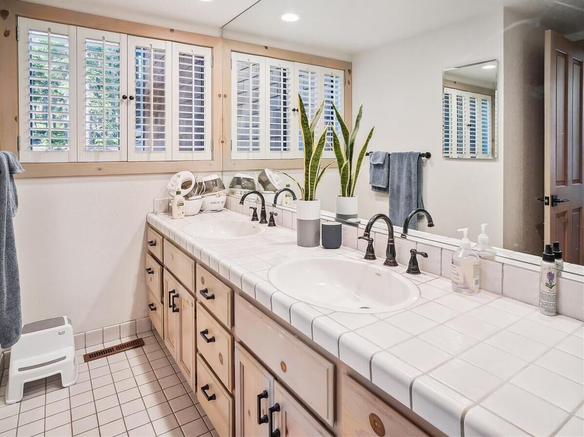 A bathroom with two sinks, a mirror, a window with blinds, and a small step stool.