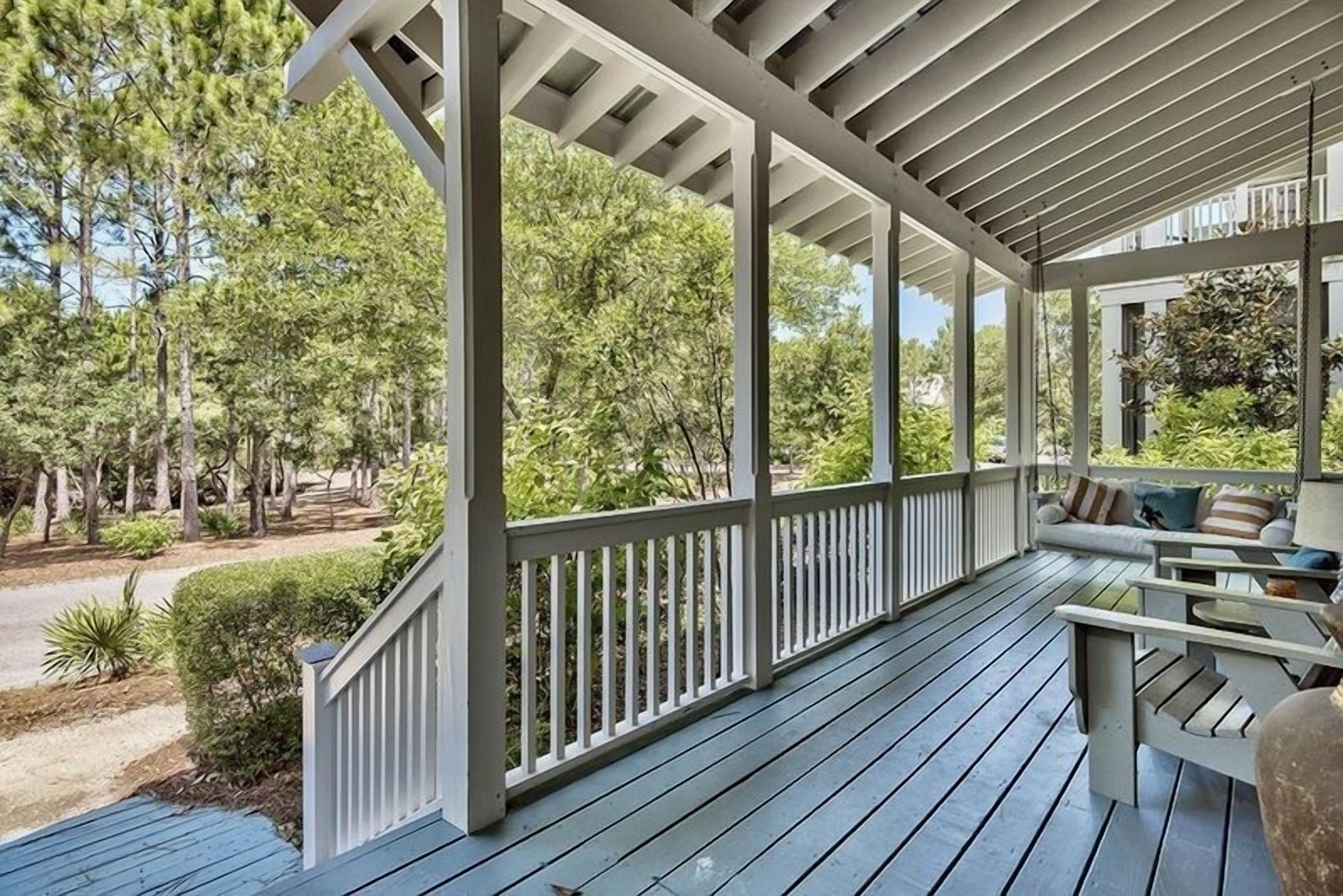 A covered porch with a wooden deck, white railings, and a seating area with cushions.