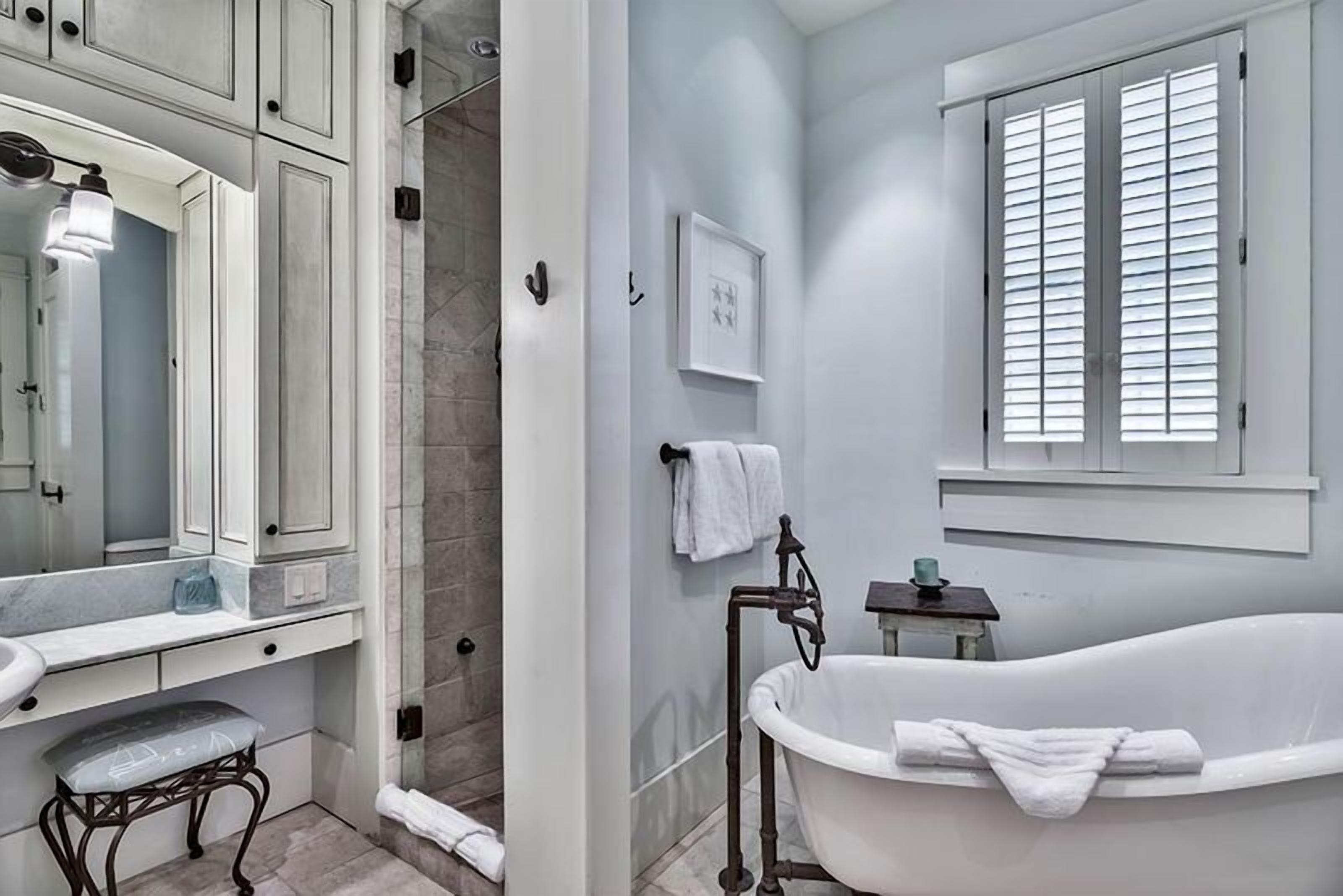 A bathroom with a white bathtub, a sink, and a mirror. There is a window with blinds and a picture frame on the wall.