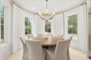 A round dining table with six chairs, a chandelier, and white curtains framing large windows. - Newly Designed Custom Seaside Home with 6 Bedrooms + Next to Adult Pool/Fitness Center 6 Home by RedAwning (Santa Rosa Beach)