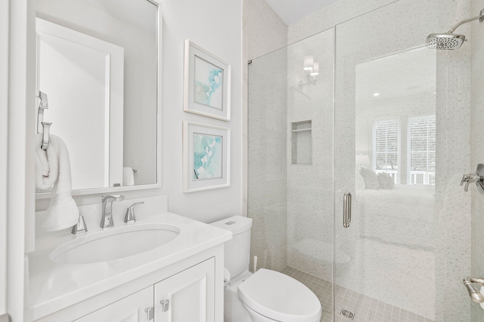 A modern bathroom with a white sink, mirror, and toilet. There are framed pictures on the wall and a glass shower enclosure.