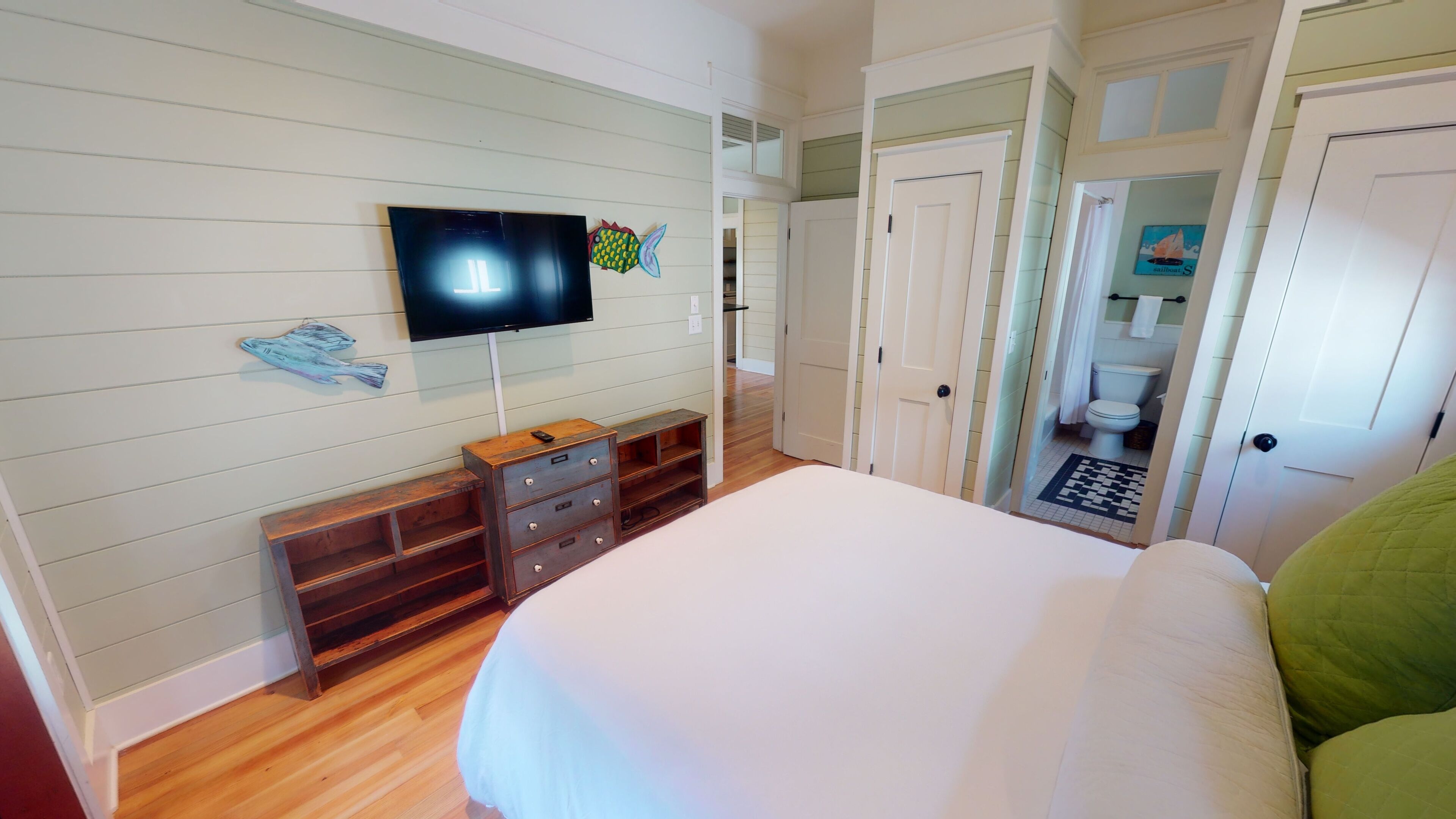 A bedroom with a bed, a TV mounted on the wall, a wooden dresser, and a door leading to a bathroom.