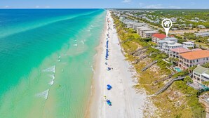 An aerial view of a beach with clear turquoise waters, a long sandy shore lined with beach umbrellas, and a row of buildings with a swimming pool and a red-roofed structure. - Shellstone Cottage Beach Front Seacrest Home with Private Pool and 5 Bedrooms 4 Home by RedAwning (Panama City Beach)