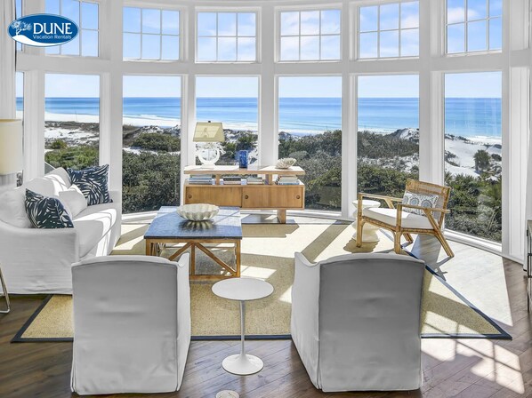 A spacious room with large windows offering ocean views, a white sofa, a dining table, and a wicker chair. - Gulf Front Penthouse Condo Complimentary Golf Cart 407 Compass Point 1, Watersound - 3 Br, Slps 8 3 Bedroom Condo by RedAwning (Santa Rosa Beach)