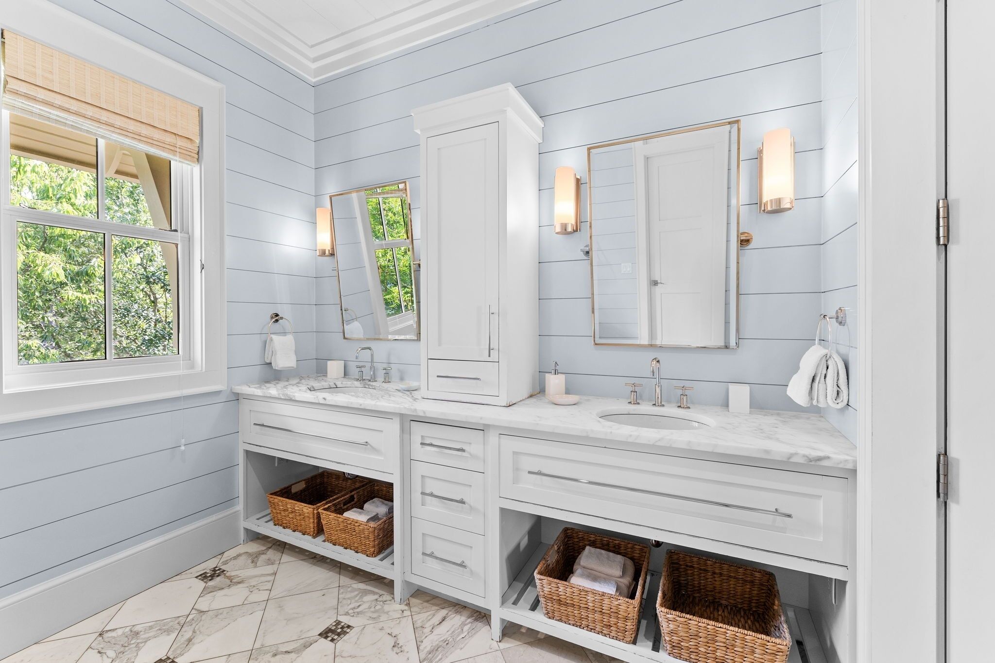 A bathroom with two sinks, white cabinetry, wicker baskets, and a window with a view of greenery.