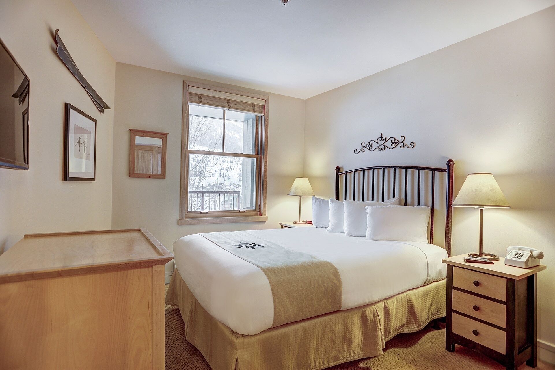 A neatly arranged bedroom with a bed, bedside lamps, a wooden nightstand, a desk, and a window with a view of trees.