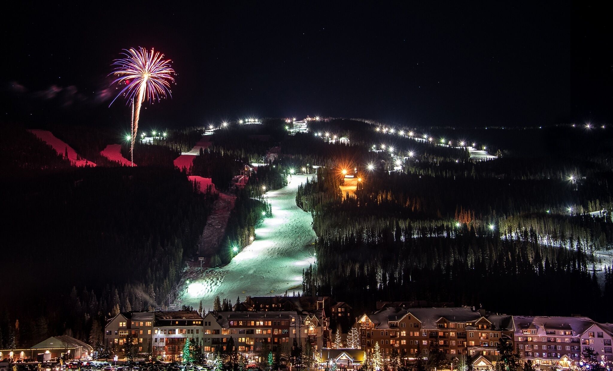Una montaña nevada con una pista de esquí, edificios iluminados y un espectáculo de fuegos artificiales.