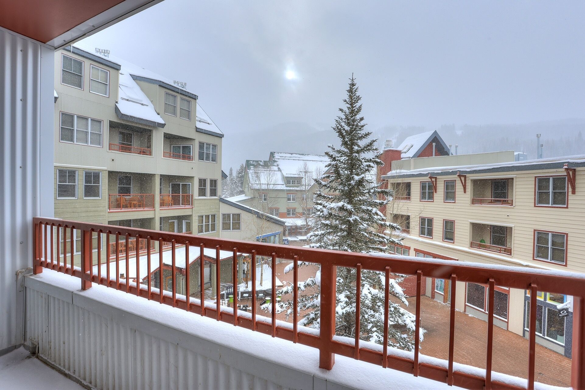 A balcony with a red railing overlooking a snowy courtyard with buildings and a snow-covered tree.