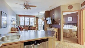A modern kitchen with a dishwasher, a dining area with a table and chairs, a ceiling fan, and a bedroom with a bed and a TV. - Silver Mill 8253-1br-walk To Slopes! Kids Ski Free! 1 Bedroom Condo by RedAwning (Keystone)