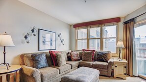 A living room with a sectional sofa, a small ottoman, a side table, and a lamp. There is a framed picture on the wall and a window with curtains. - Silver Mill 8253-1br-walk To Slopes! Kids Ski Free! 1 Bedroom Condo by RedAwning (Keystone)