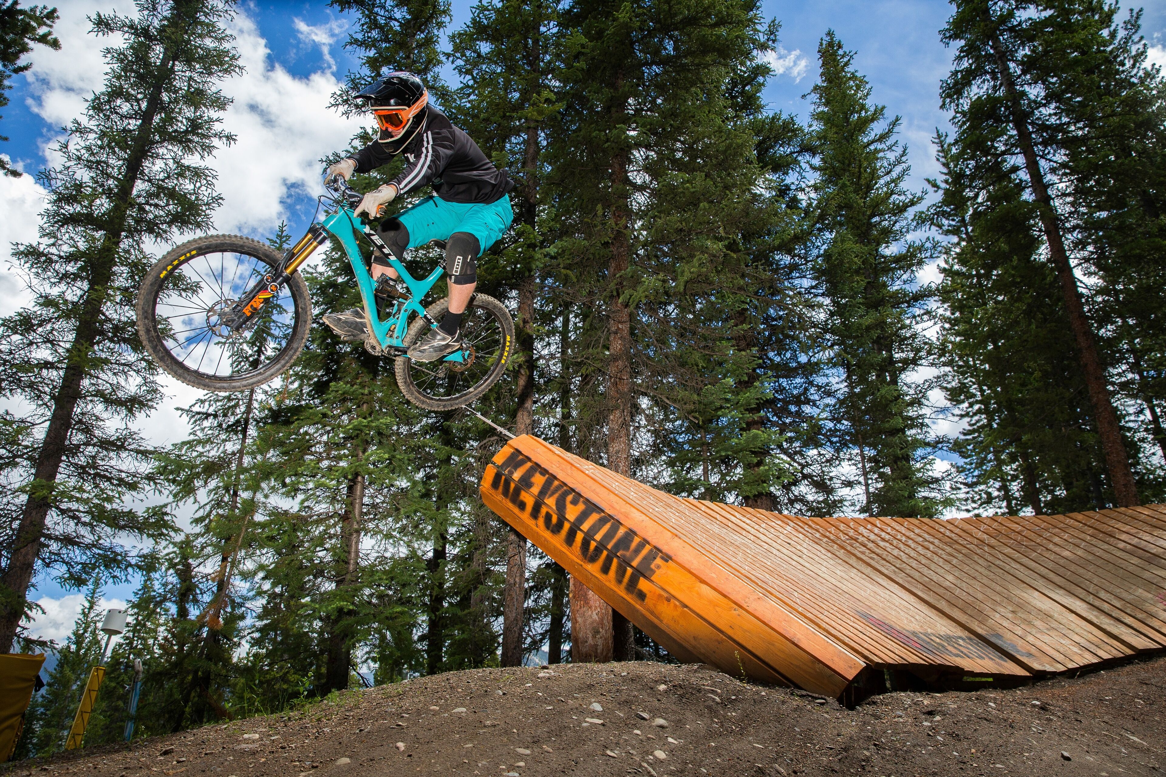 A mountain biker performing a jump over a wooden ramp with the word "REXSTONE" on it.