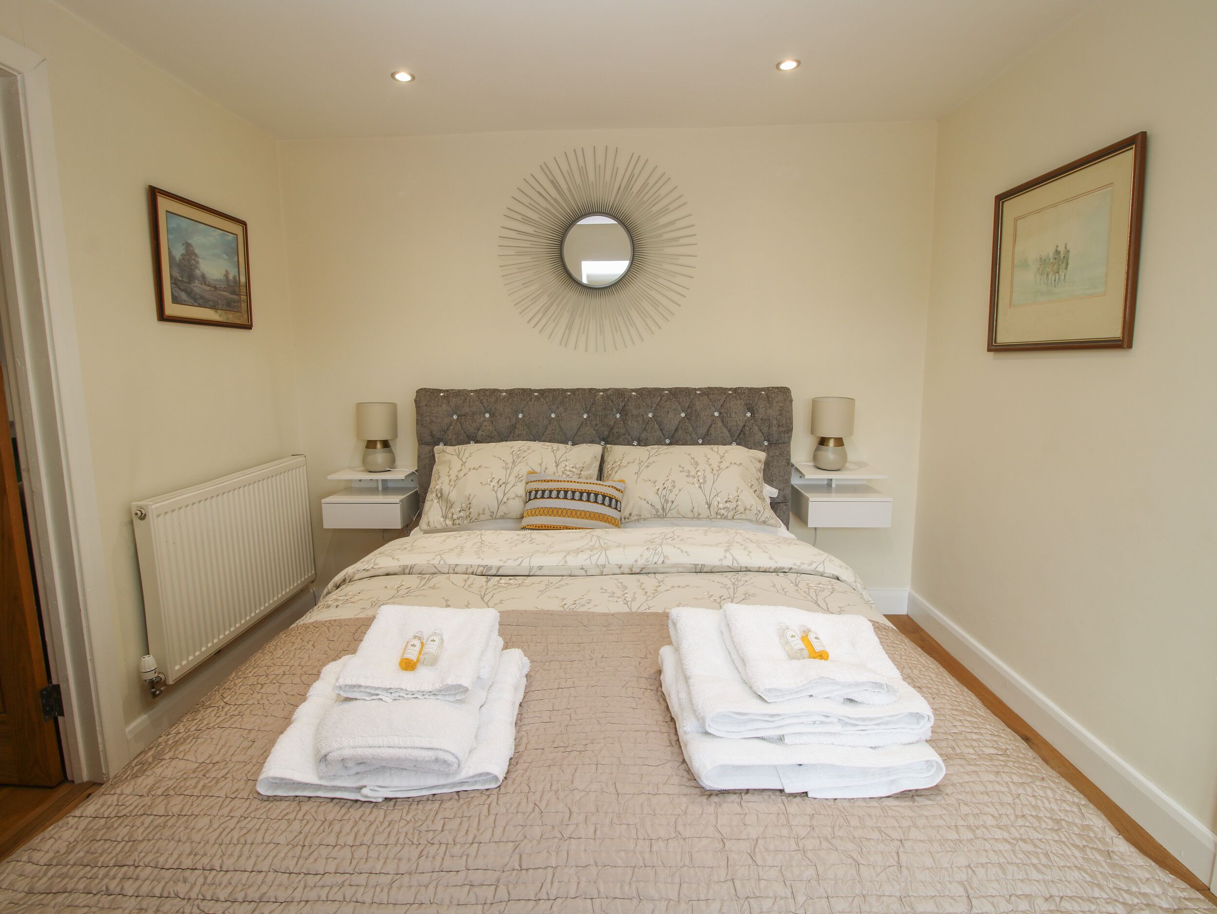 A neatly made bed with a tufted headboard, two bedside tables with lamps, a radiator, and two framed pictures on the walls.