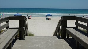 Una pasarela de madera que lleva a una playa con dos sombrillas azules y algunas personas a lo lejos.
