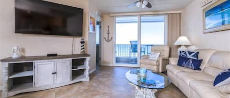 Una sala de estar con televisor de pantalla plana, ventilador de techo, una mesa de centro de vidrio, sofá color beige y balcón con vistas.