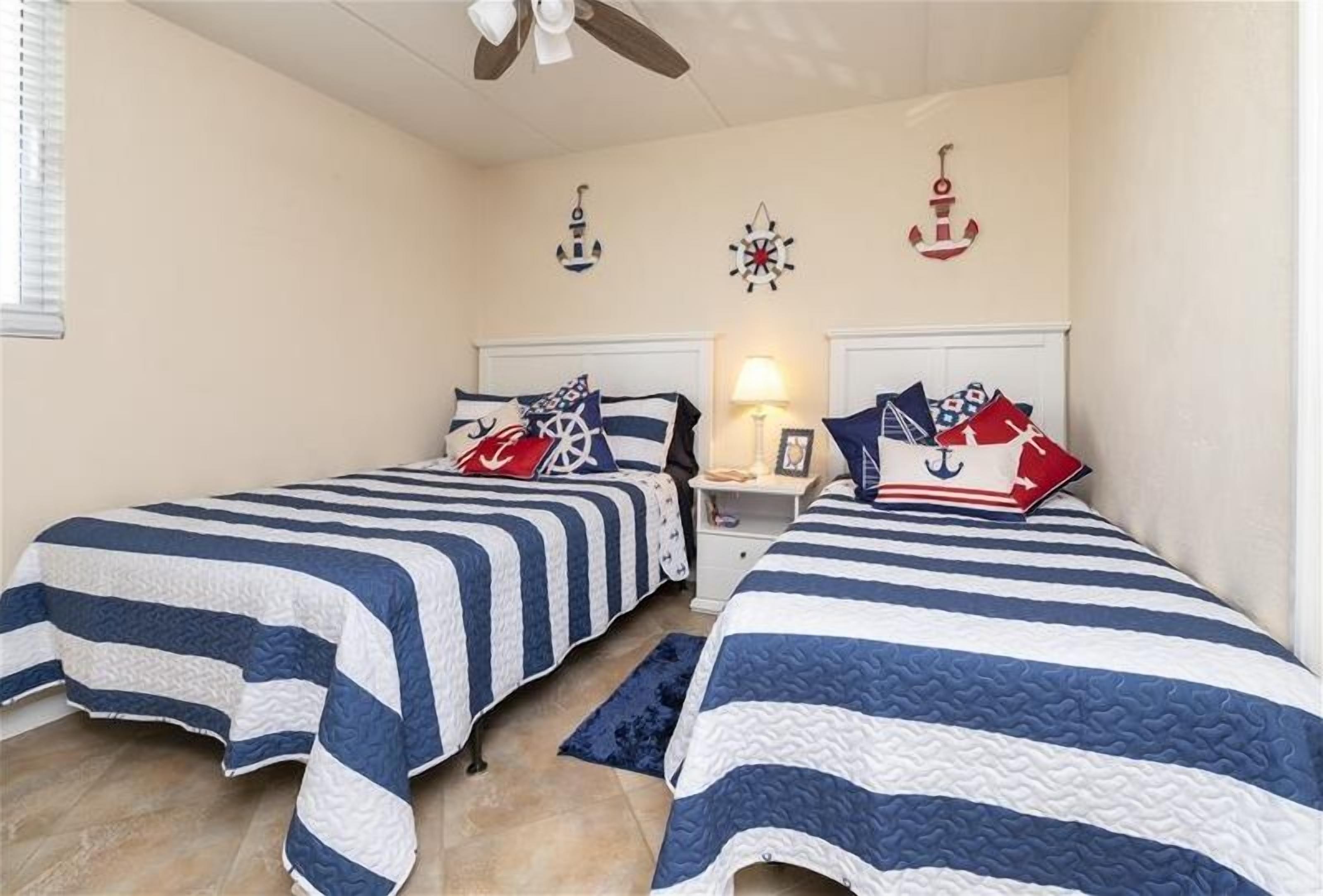 A bedroom with two beds, each with striped bedding, a ceiling fan, and nautical-themed wall decorations.