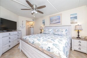 Um quarto com uma cama, um ventilador de teto, uma televisão, uma cômoda e uma mesa de cabeceira.