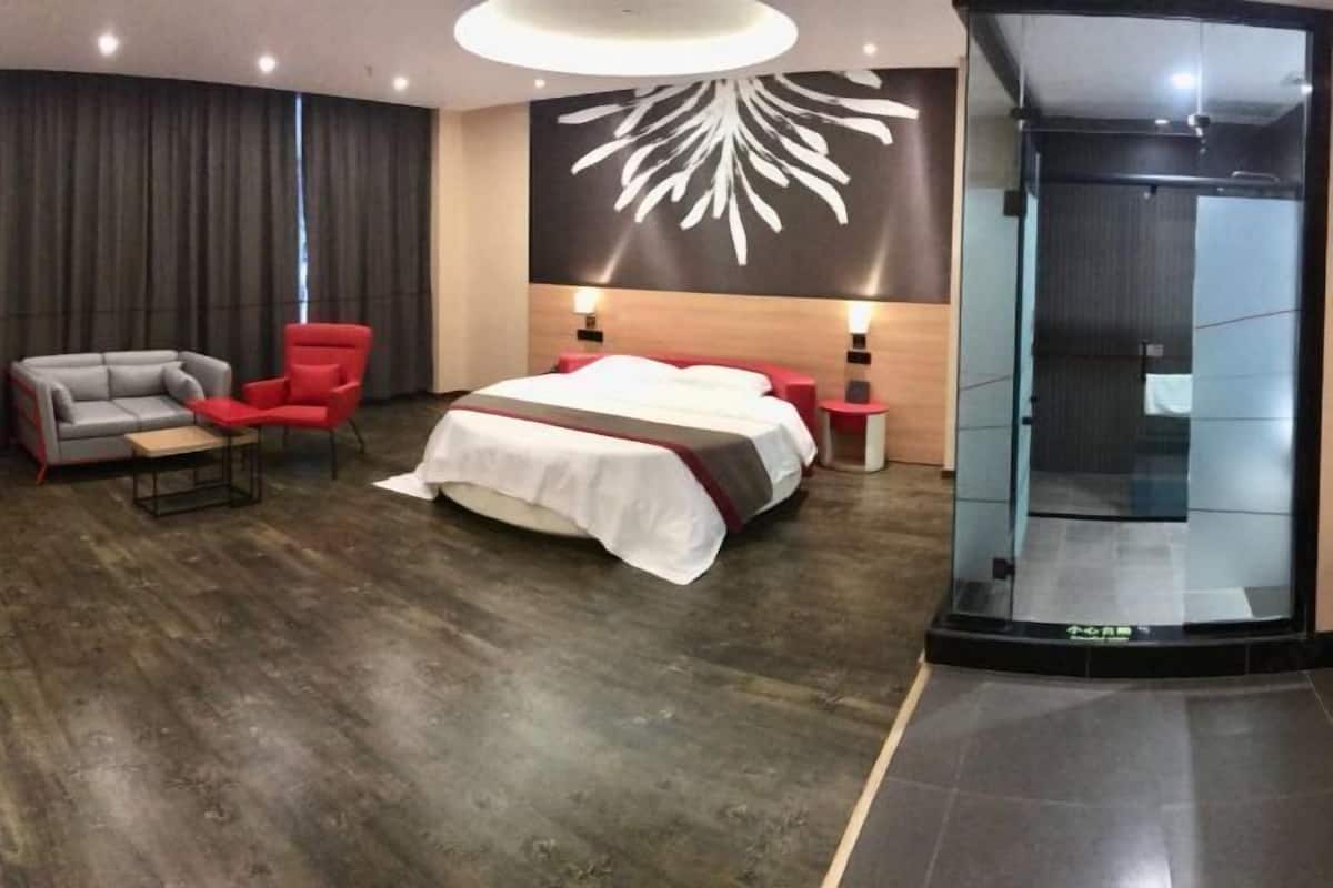 Ein modernes Hotelzimmer mit einem großen Bett, einem roten Sessel, einem grauen Sofa und einem Glaselevator.