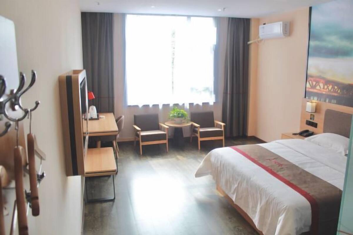 Ein Hotelzimmer mit einem Bett, einem Schreibtisch mit Stuhl, einem kleinen Tisch mit zwei Stühlen, einem Fernseher und einem Fenster mit Vorhängen.