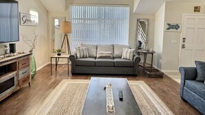 Ein Wohnzimmer mit einer Couch, einem Couchtisch und einem Fernseher. Es gibt ein Fenster mit Jalousien, eine Lampe und ein Schild mit der Aufschrift "Amazing Grace".