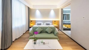A modern hotel room with a green sofa, a white coffee table with a vase of flowers, a bed with white linens, and a bathroom visible through an open door. - Guangyi Hotel (Quanzhou)